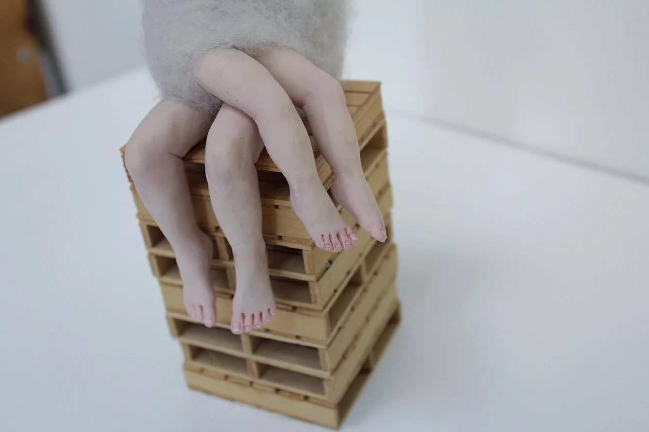 Doll with realistic skin, hanging off a stack of wooden pallets on a white surface.