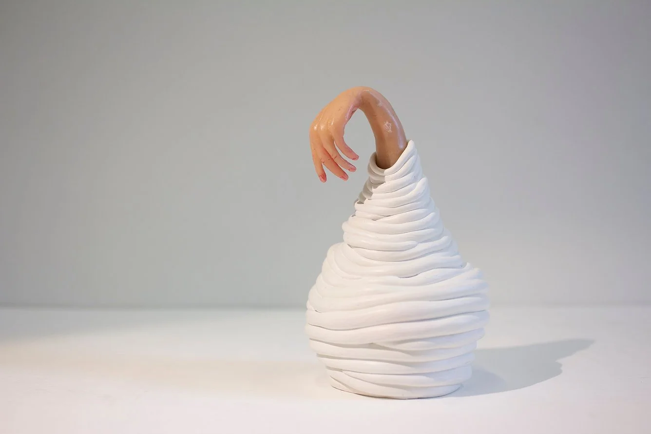 A white twisted ceramic-like vase with a realistic human hand sculpture emerging from the top.