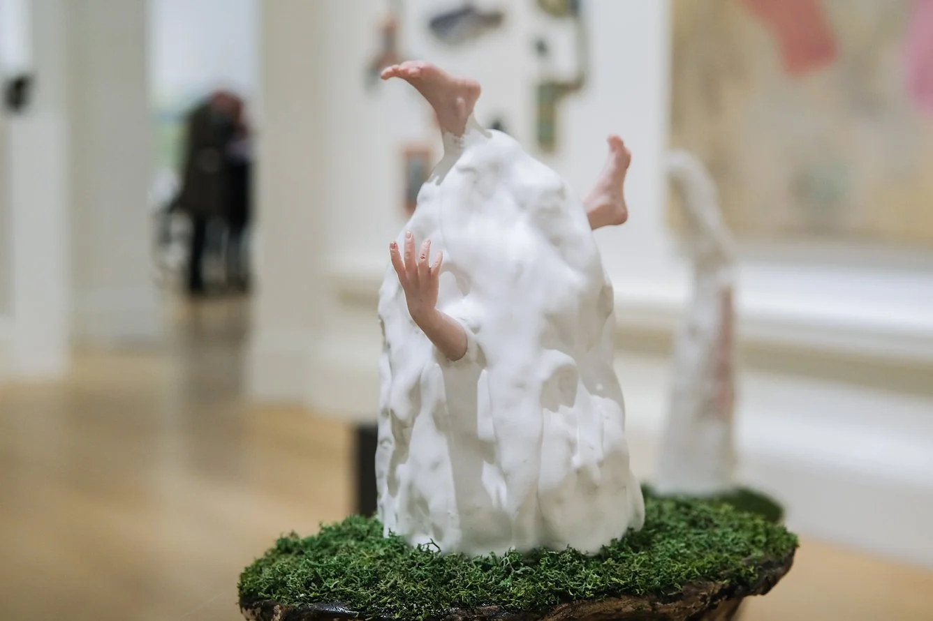 Contemporary sculpture of a person's head and hands emerging from a white, textured material on a mossy base, displayed in an art gallery.