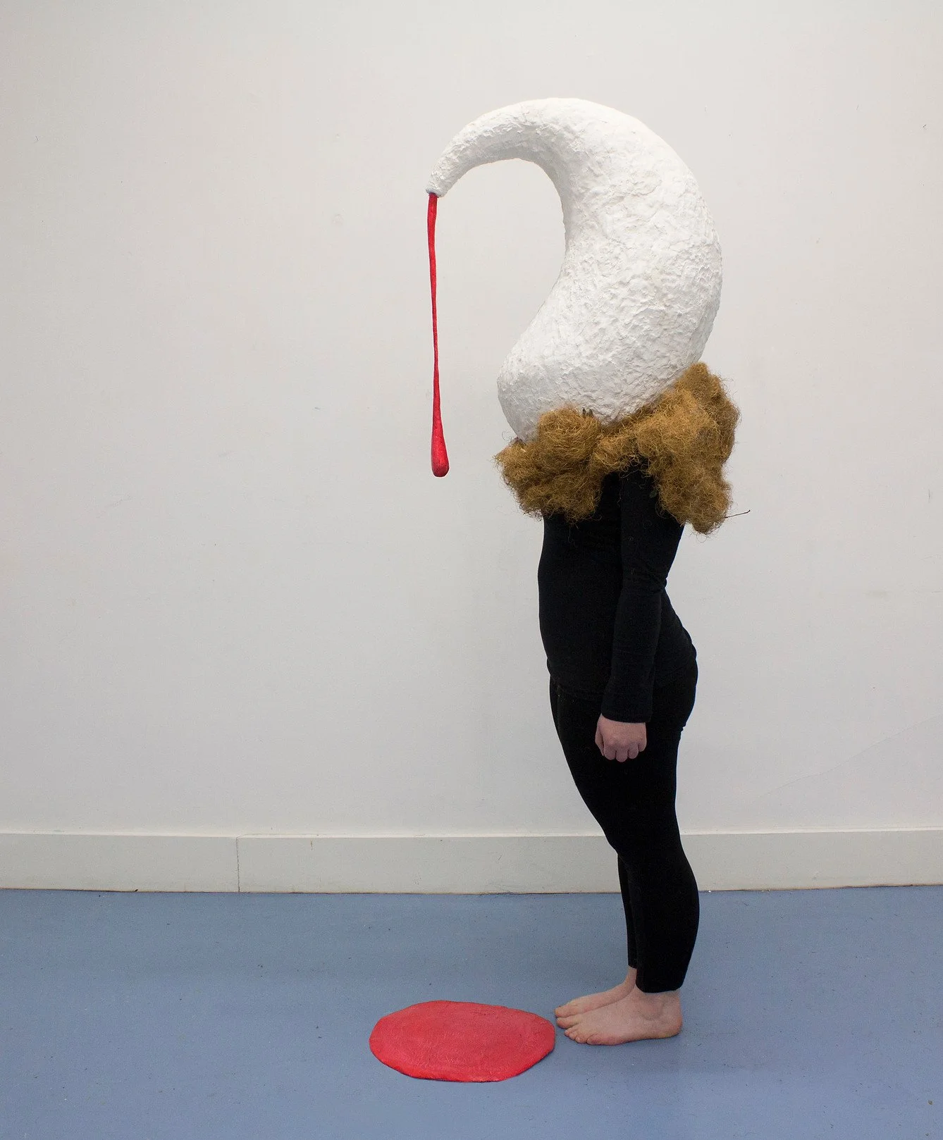 Person wearing a large  swan-like mask head with a red dripping beak, black clothes, and a brown fuzzy collar, standing on a blue floor with a red circular mat in front, against a plain white wall.