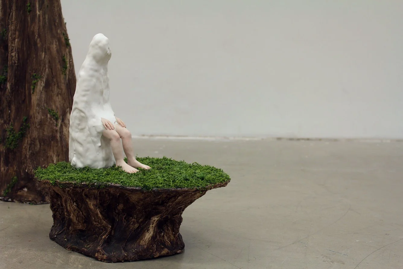 A sculpture of a faceless, featureless figure sitting on a moss-covered wooden platform next to a tree trunk, with a plain background.