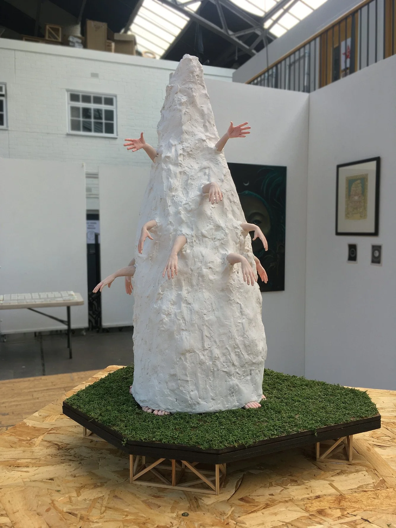 Sculpture resembling a tall white mountain with multiple human arms protruding from its sides and top, displayed on a small platform covered with green moss, in an art gallery setting.