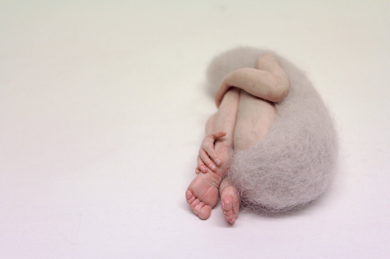 Close-up of a handmade felted sculpture of a sleeping figure curled up on its side.