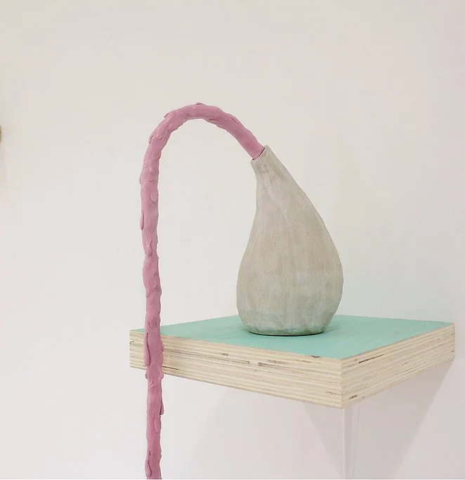 A ceramic teardrop-shaped vase with a long pink form spurting out of the spout and aqua-colored platform.