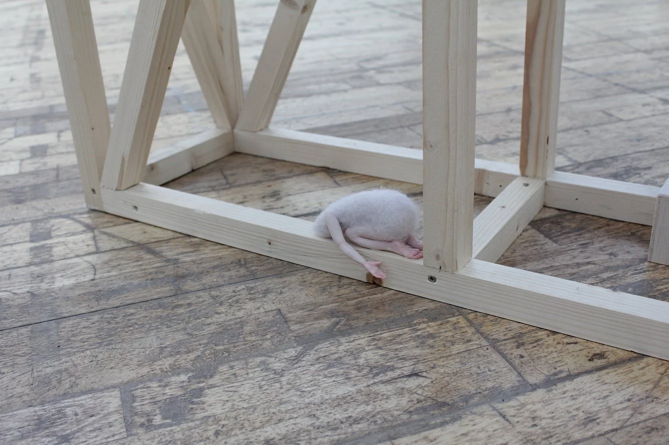A small felted figurine, resting beneath a wooden structure on a wooden floor.