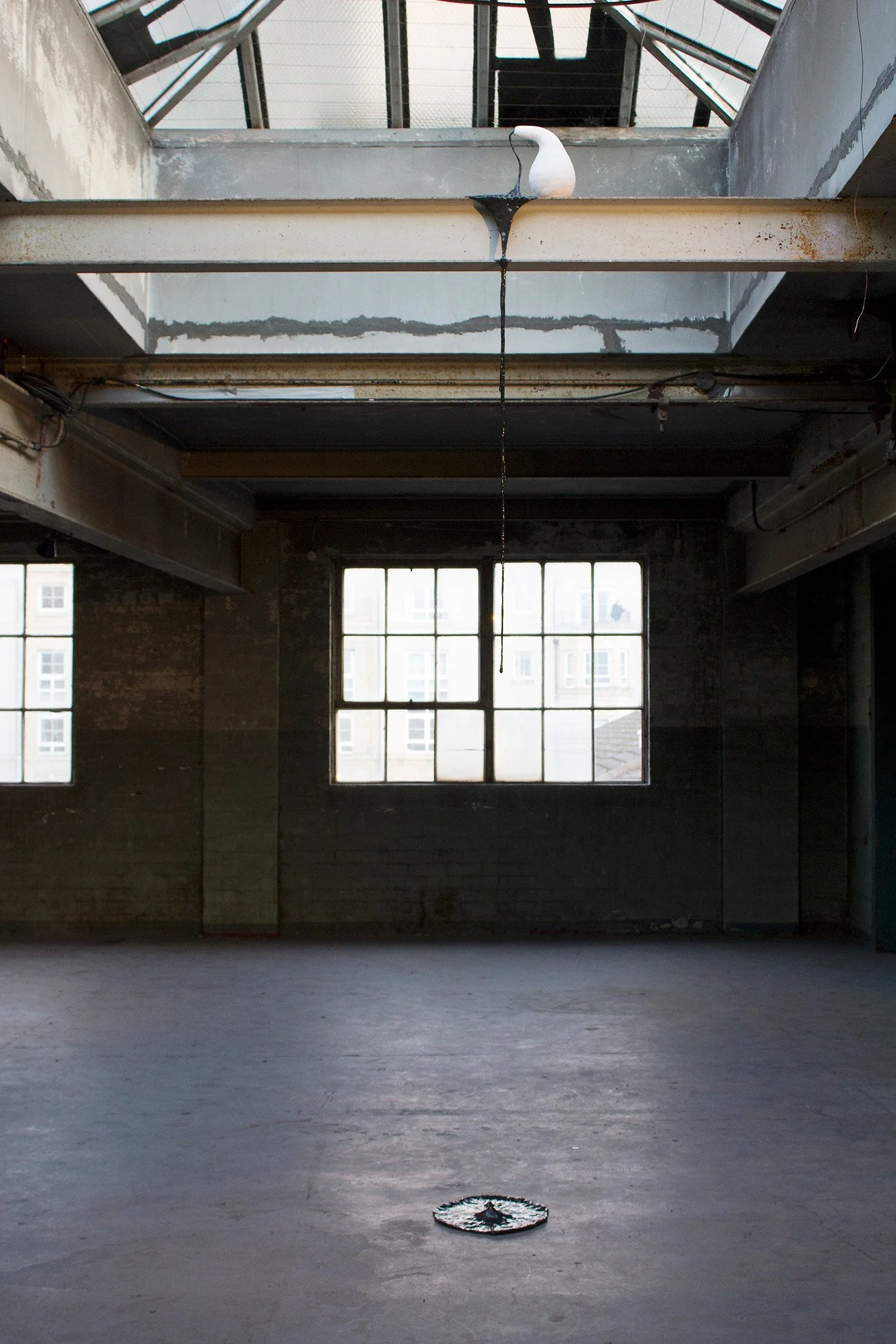 An industrial-style room with large windows and a glass ceiling, featuring a white ceramic sculpture mounted on a metal beam and a circular object with a star in the center on the floor.