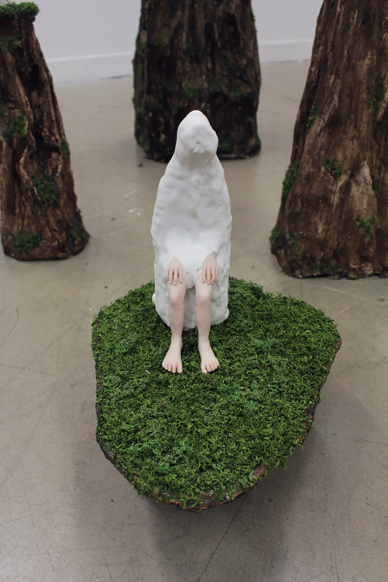 An art sculpture featuring a humanoid figure with a featureless white head and hands, sitting on a moss-covered platform, surrounded by tree trunks.