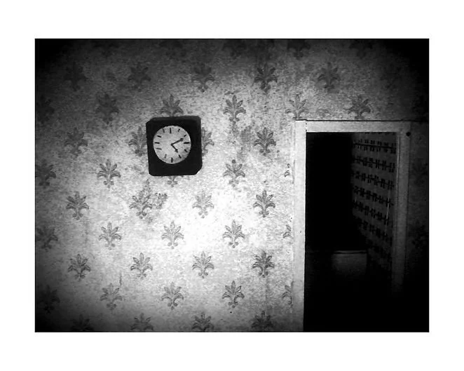 Black and white photo of an interior wall with fleur-de-lis patterned wallpaper, a clock showing approximately 4:10, and an open doorway leading to a dark room with shelves or mailboxes.
