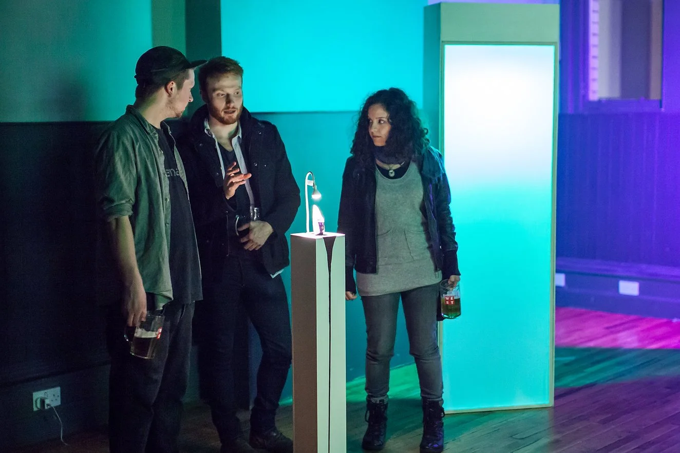 Three young adults in a dimly lit indoor space, standing near a small display with a lit lamp and a bottle, illuminated by colorful lighting, engaged in conversation.