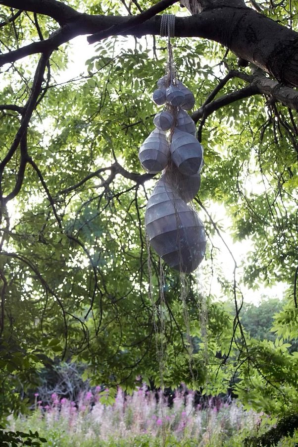 Hanging art installation with several metallic, conical shapes in a tree with green foliage and a field of pink and white flowers in the background.