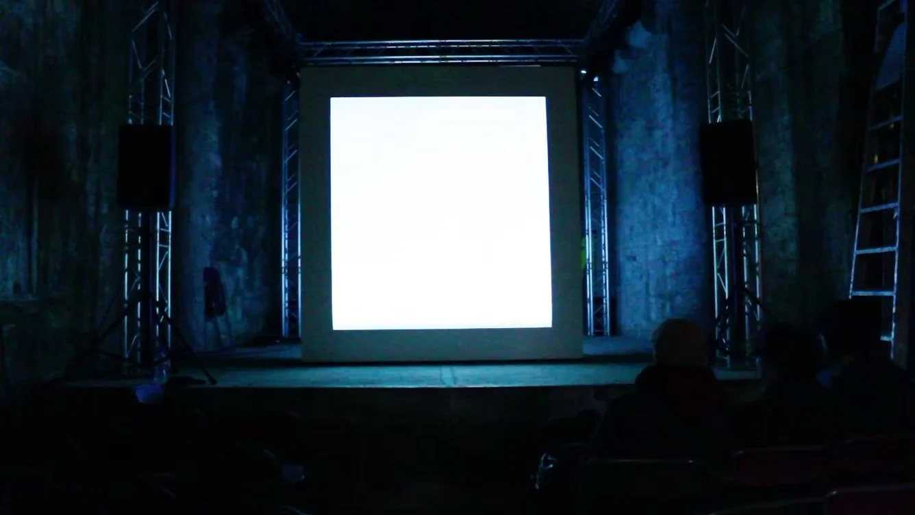 A stage setup with a large illuminated screen, speaker stands, and audience members sitting in a dark room.