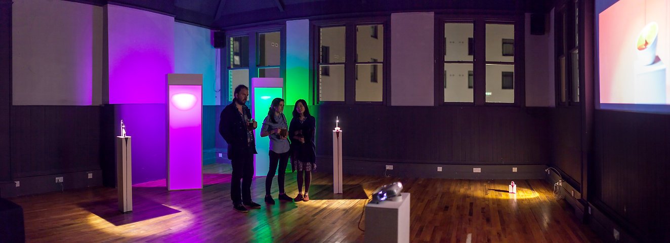 Three people stand in a dimly lit art gallery or exhibition space with colorful lighting and modern display pedestals.