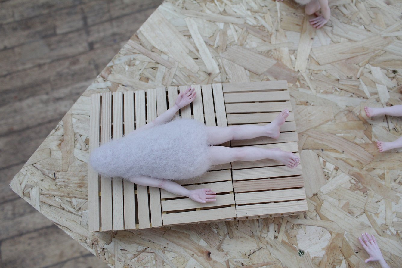 A small figurine of a person lying face down on a wooden slatted platform, surrounded by other tiny doll-like feet and hands on a wooden surface.