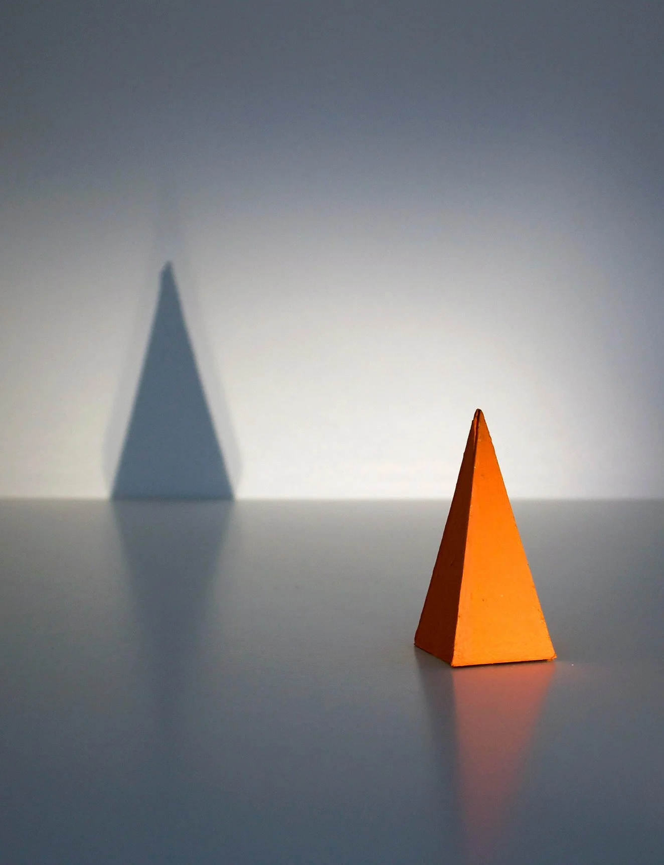 An orange pyramid-shaped object casting a shadow with a distinct dark blue pyramid-shaped shadow on the wall behind it.