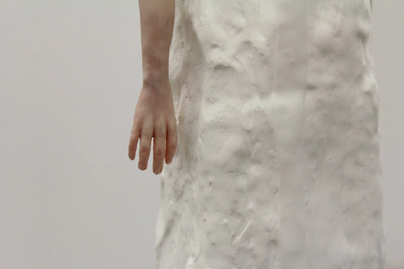 Close-up of a person’s hand and part of their white textured clothing against a plain background.