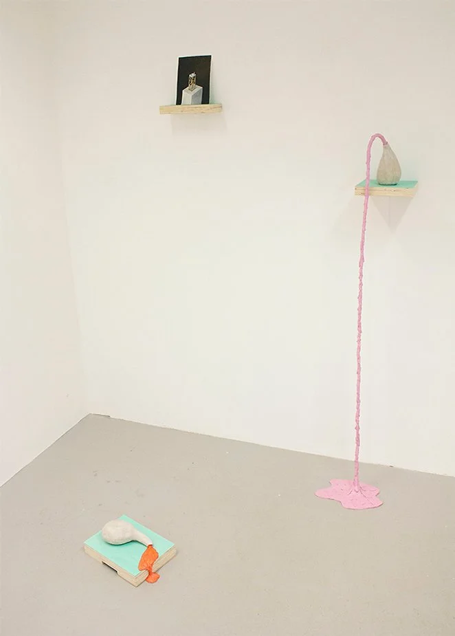 Minimalist art installation with small shelves holding objects, a pink thread hanging from a shelf to the floor with pink fabric pooled on the ground, and a woven piece with a round shape and a small orange decoration on a wooden platform.