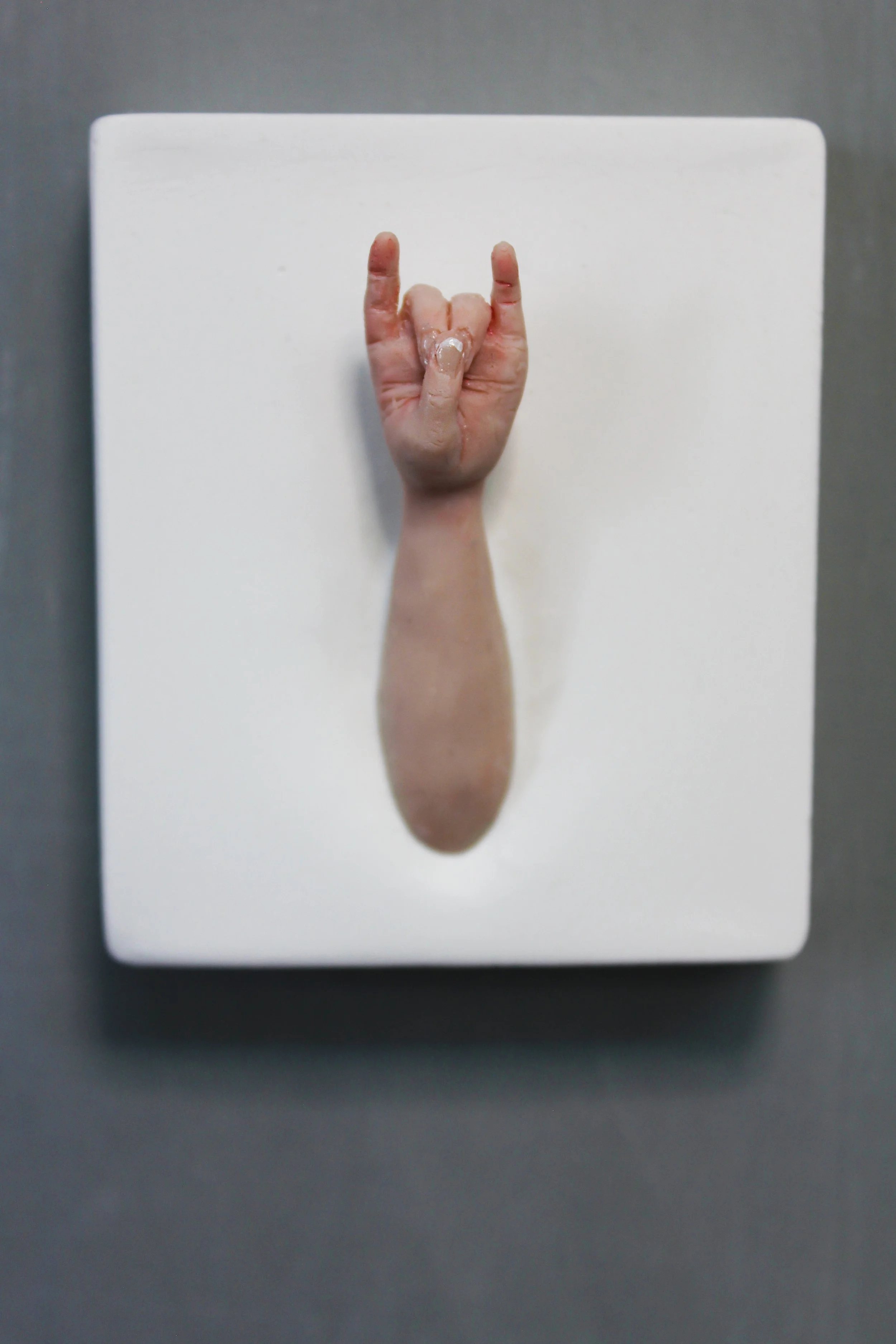 A close-up of a white textured surface with a realistic looking sculpture of a human hand making a 'rock on' gesture, with index and pinky fingers raised and thumb holding down middle and ring fingers.