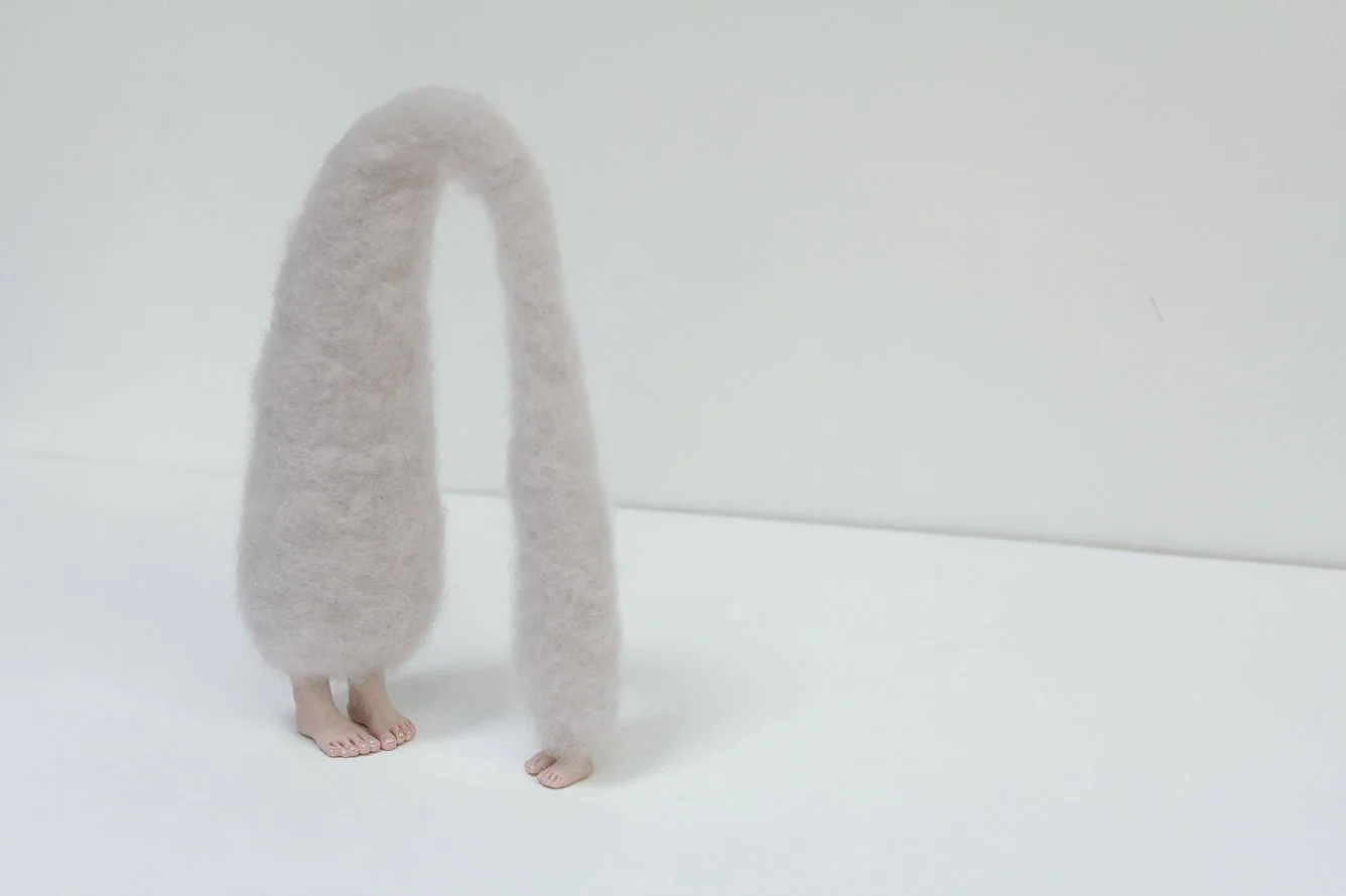 A person with pink skin and bare feet is covered from the torso up by a large fluffy, light-colored furry object, creating an abstract form against a plain white background.
