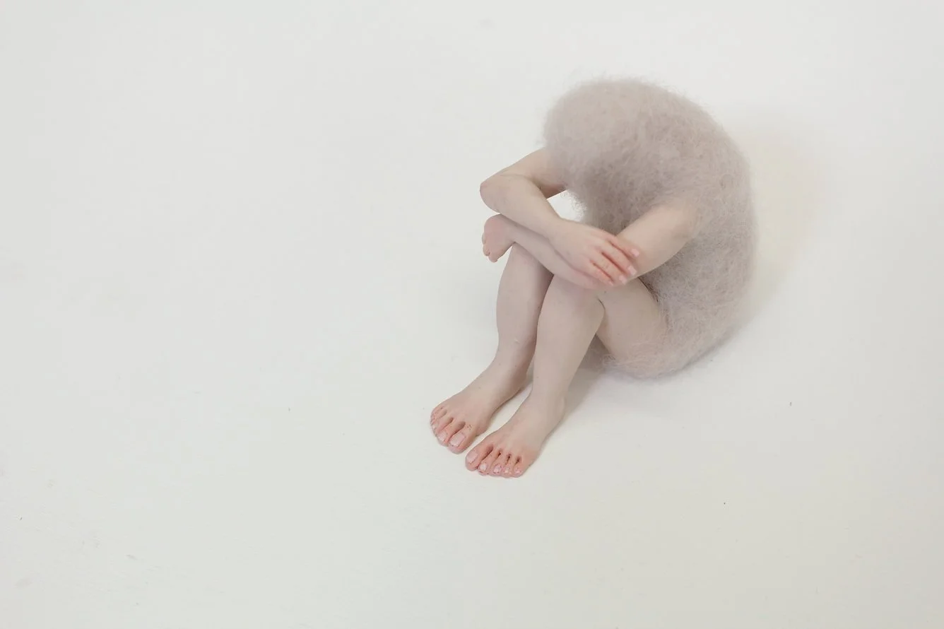 A felted figurine sitting with knees drawn up and arms wrapped around their legs, blending into a white background.