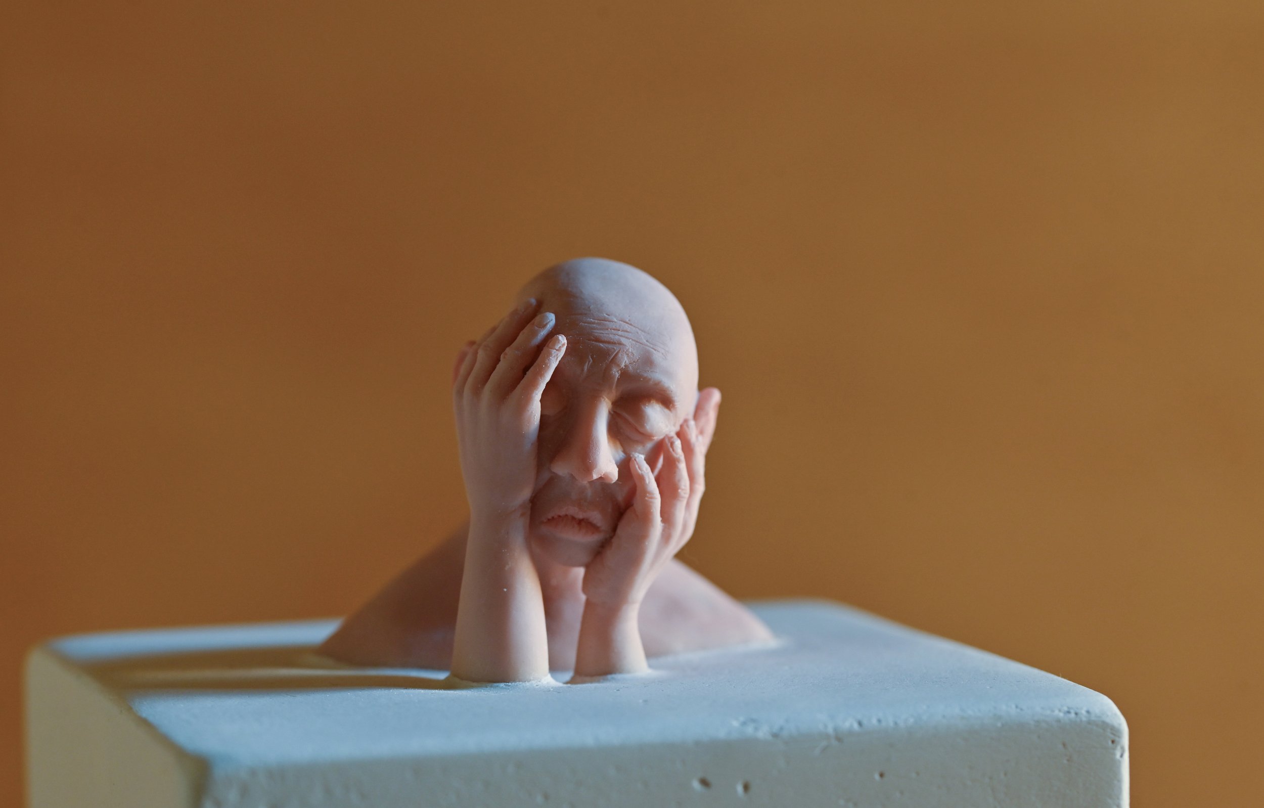 A sculpture of a person's head and hands, with hands covering part of the face, placed on a pedestal against a plain brown background.