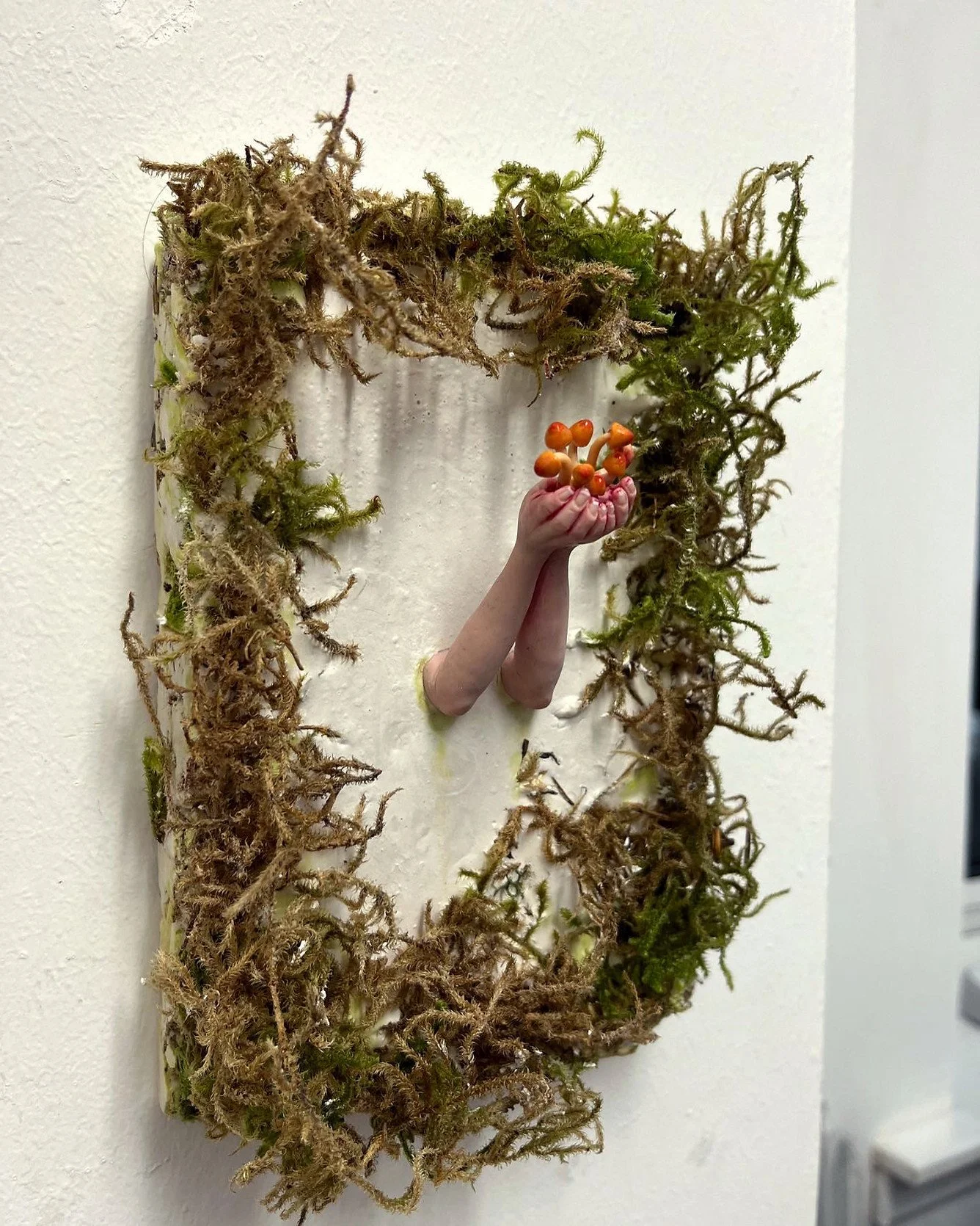 An art installation resembling a mirror frame made of moss and twigs, with a human arm and hand emerging from it holding a bunch of small orange fungi.
