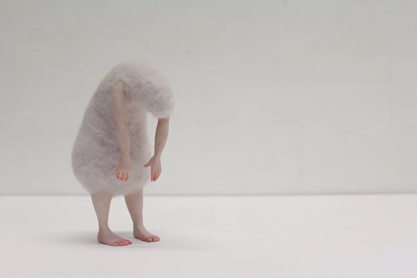A surreal sculpture of a person with a head covered in a fluffy white material, bent over with arms hanging down, standing on a plain white surface against a plain white background.