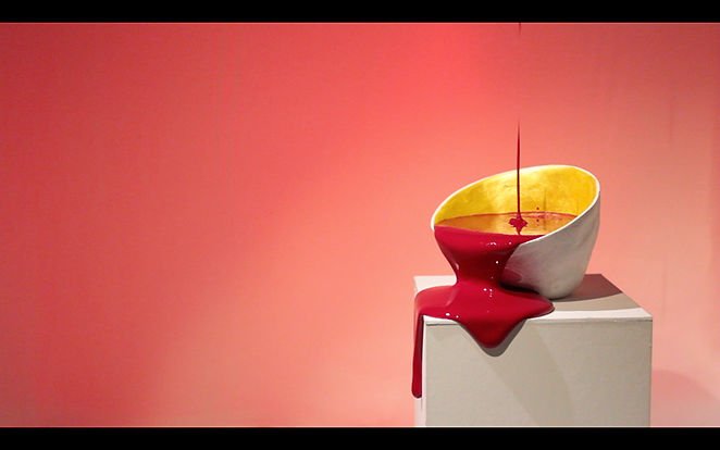 A modern art sculpture resembling a melting bowl of yellow and gray with red liquid spilling over the edge, placed on a white pedestal against a pink background.