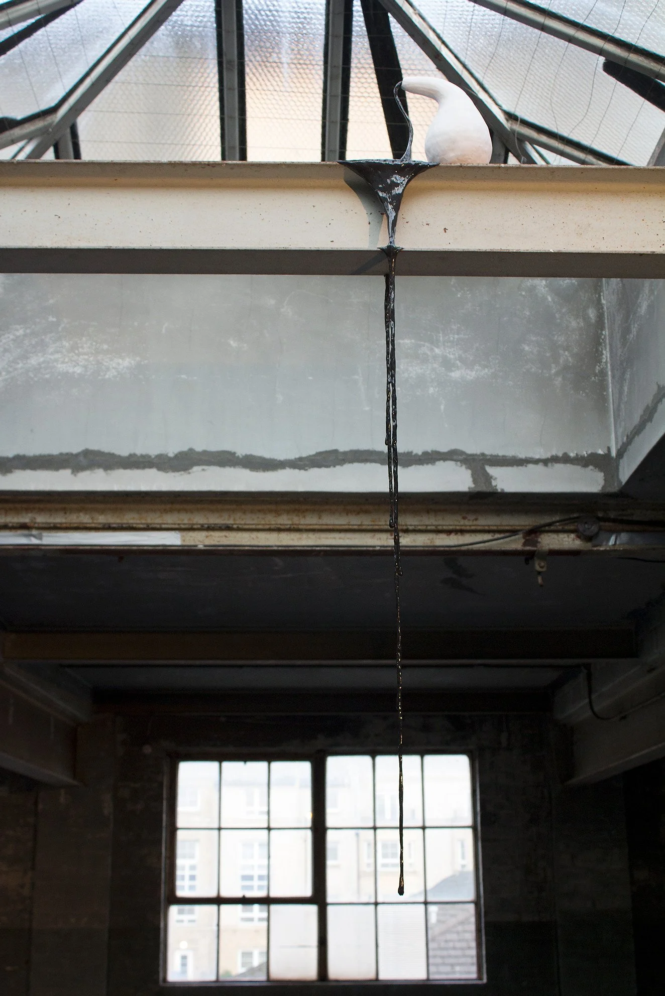 A white ceramic pot standing on the edge metal beam, with melted black wax dripping down from the top edge of the case, inside a building with large windows.