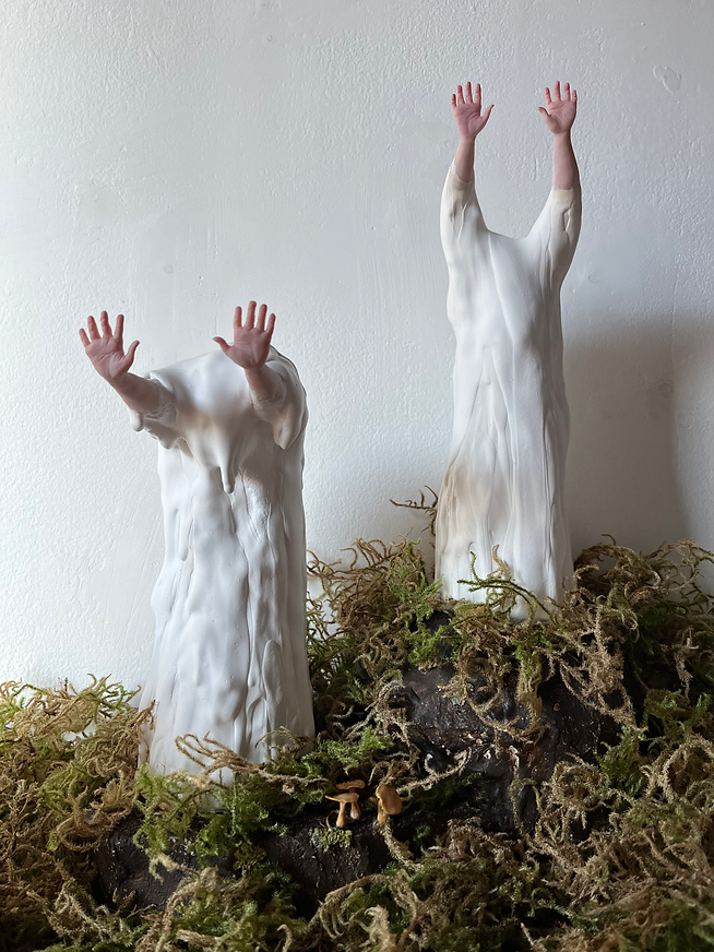 Artistic sculptures of two figures with elongated bodies and raised arms, surrounded by moss and small mushrooms, against a plain white wall.