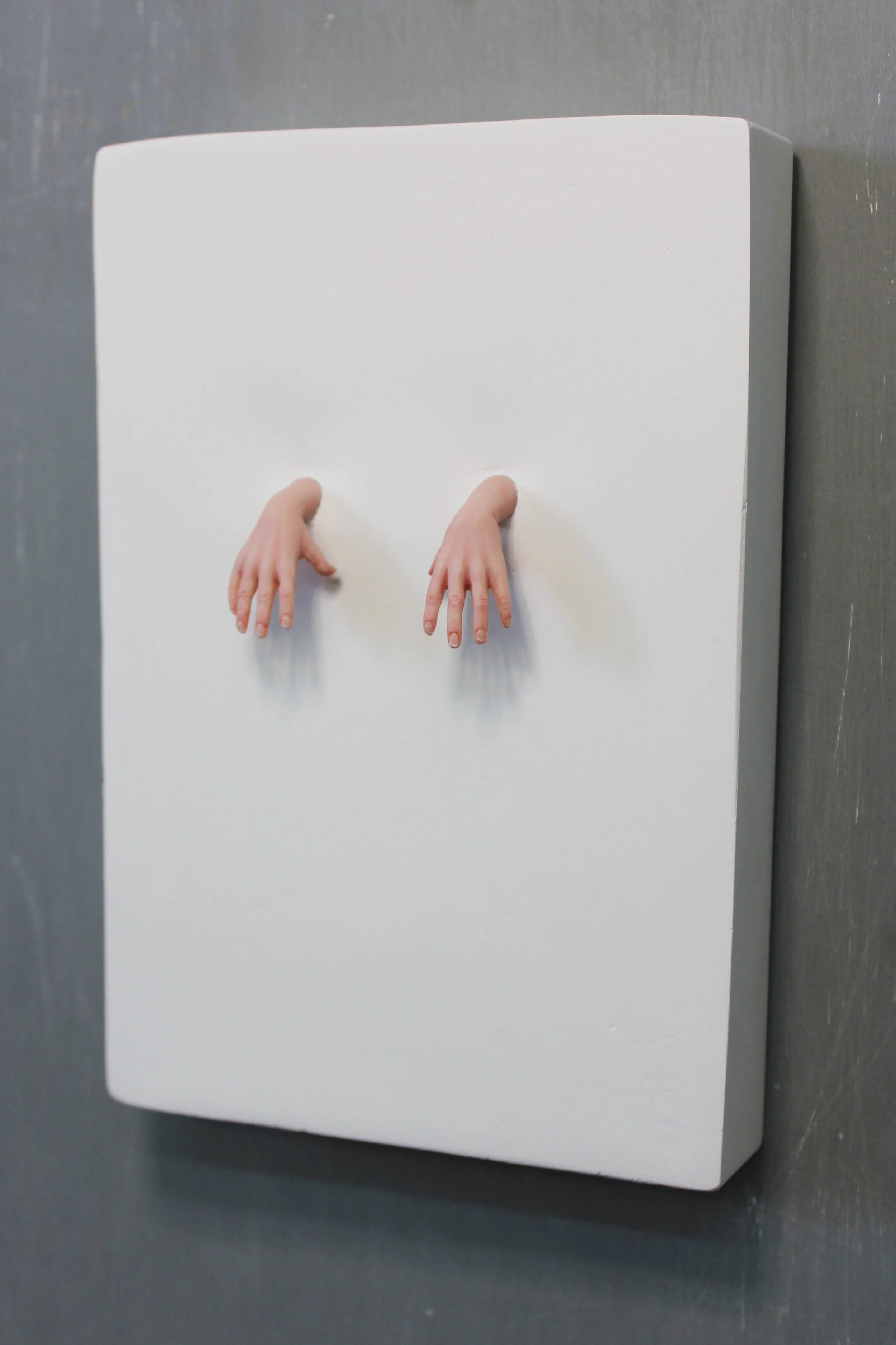 An art installation on a wall featuring two realistic sculpted children's hands protruding from a white panel.