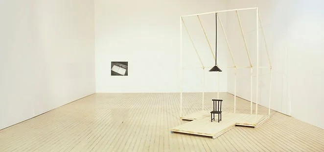 An empty art gallery with a wooden floor and white walls, featuring a minimalist sculpture of a small black chair on a tilted wooden platform and a swing hanging from a white frame.