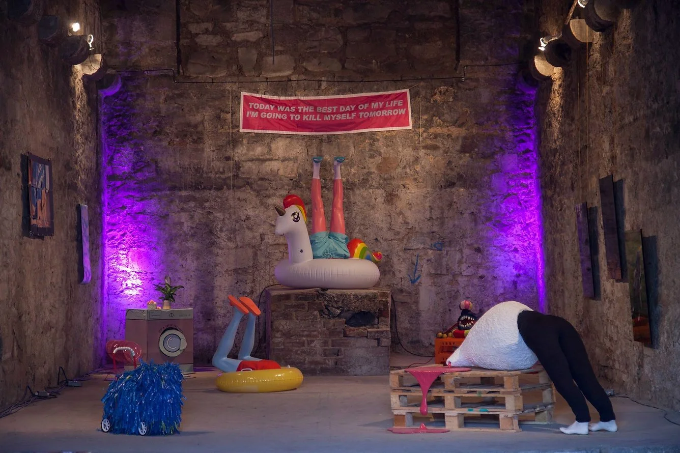 An art installation in a brick-walled room features a person with a white, textured head and black pants bending over, with colorful inflatable objects, a small washing machine, a blue pom-pom covered skateboard, and a pink banner with text. The bann