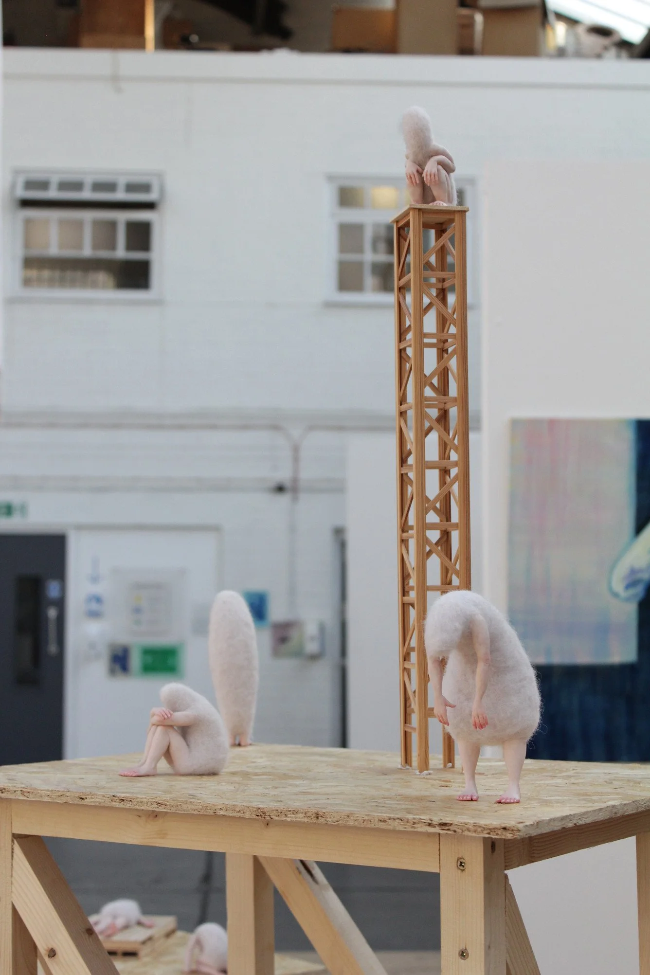Miniature woolly  figures are positioned on a wooden table, with one climbing a lattice tower, one standing upright, and one sitting with knees drawn up. The background shows a white industrial wall with windows.