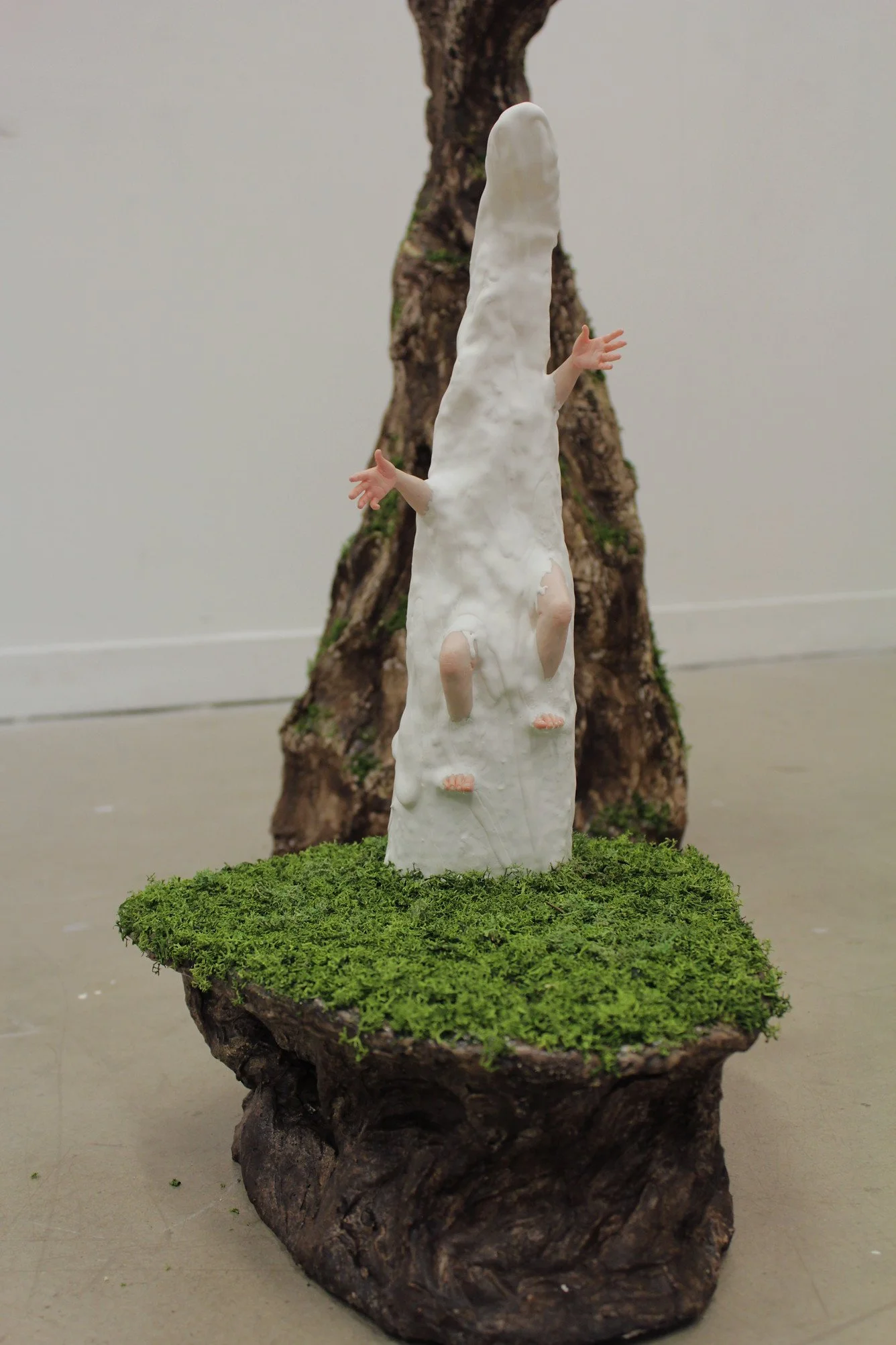 A sculpture featuring a face-down human figure with arms outstretched, embedded in a tall, white, textured structure resembling a tower, set on a moss-covered base in front of a piece of bark or wood.