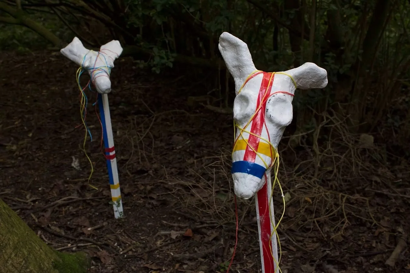 Two white horse-shaped sticks decorated with colorful tape and yarn, placed in a wooded outdoor area.