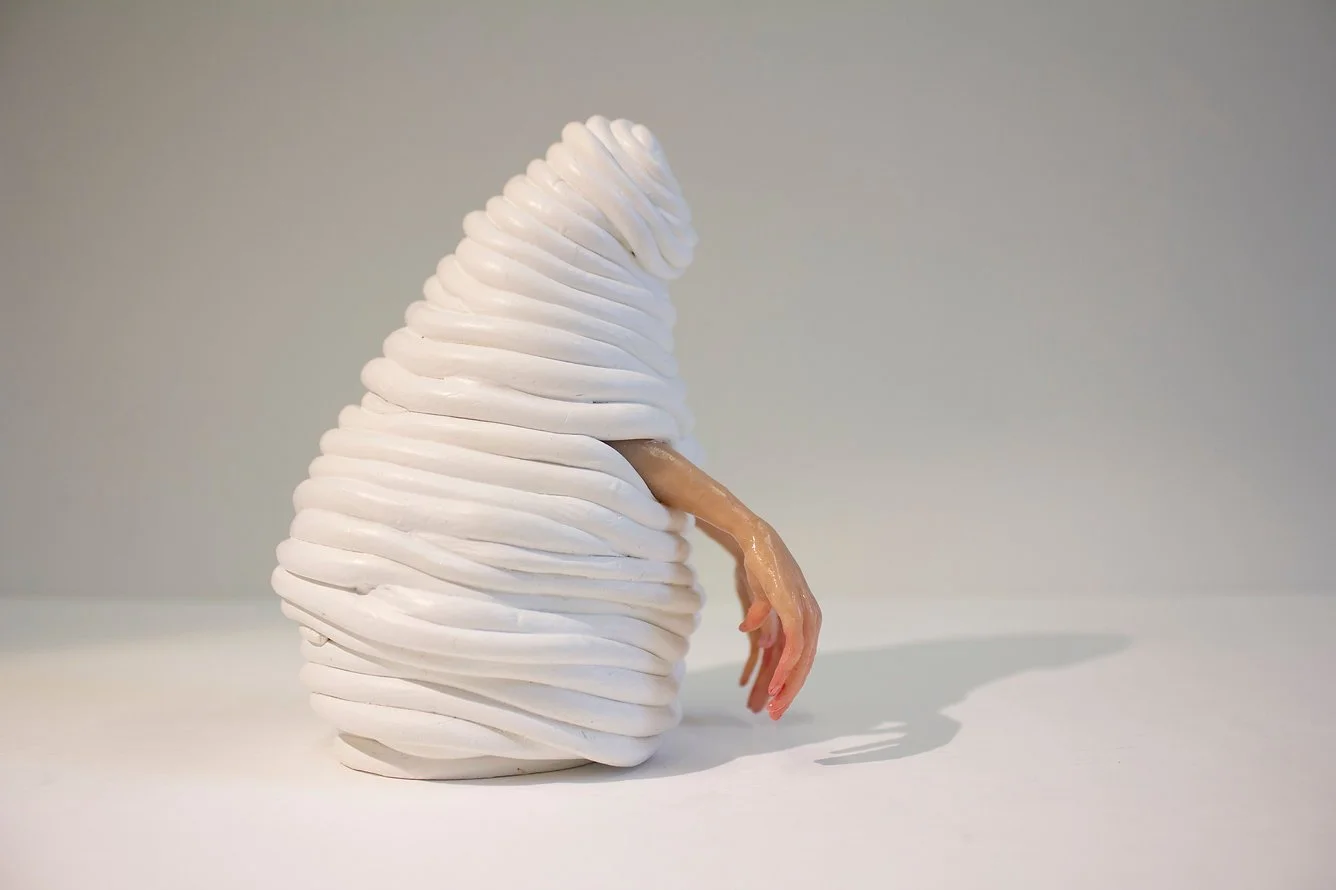 An abstract sculpture resembling a pear made of white layered material with a sculpted human arm hanging from it, cast in a peachy color.