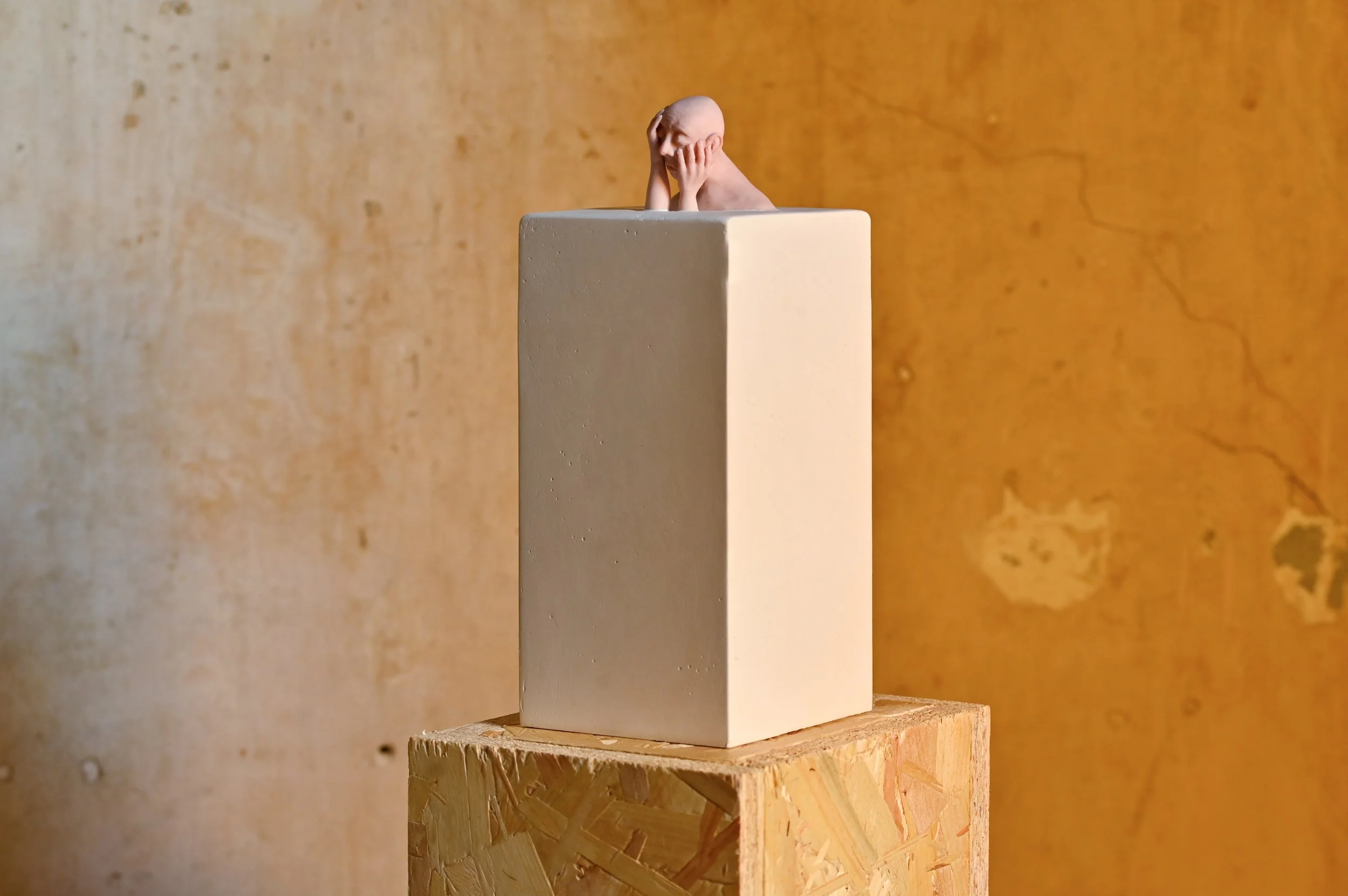 Small sculpture of a human head with hands on cheeks, placed on a white rectangular block, set on a wooden stand against a textured orange-brown background.