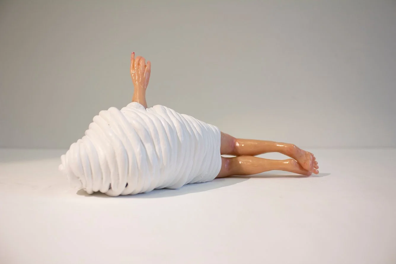 A sculpture of a human figure lying on its side made from whipped cream-like polymer clay and plastic parts, with the head and arms raised.