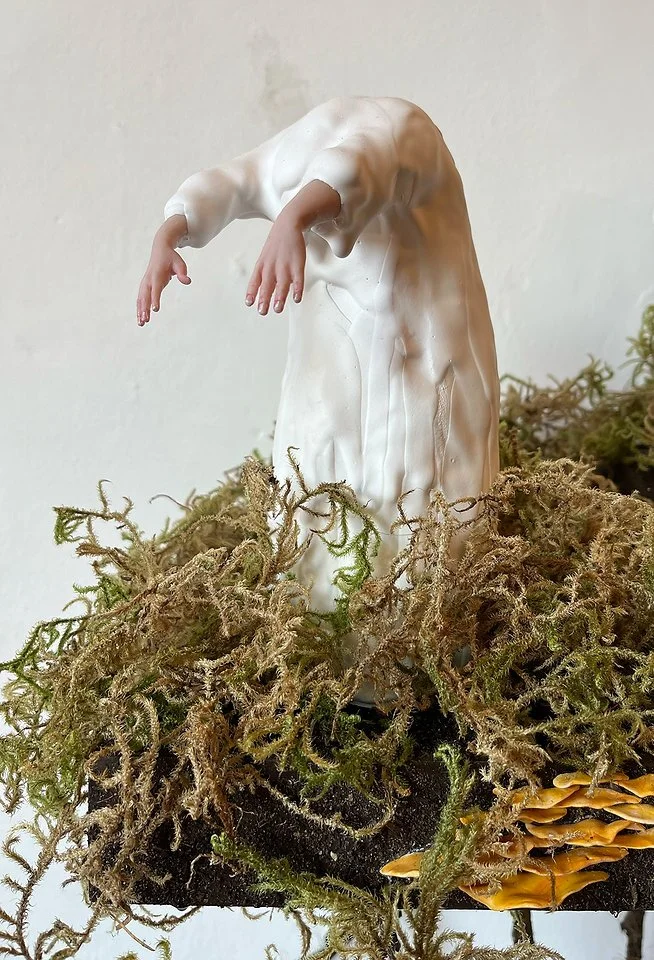 A small white sculpture of a person with arms hanging down, standing on a base surrounded by dried moss and fungi, against a plain white wall background.