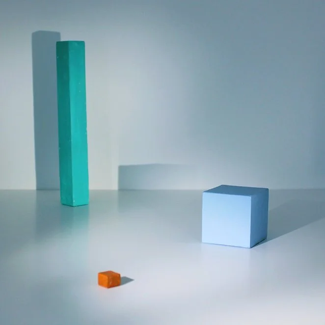 Three geometric blocks of different sizes and colors sit on a white surface with shadows cast behind them. The blocks are turquoise, white, and orange, with the tallest turquoise block on the left, a large white block on the right, and a small orange