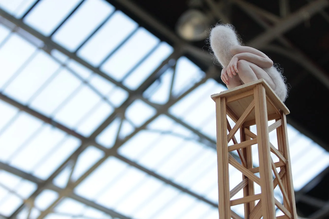 A small figurine of a person , sitting with knees pulled up to chest,  perched on top of a wooden structure resembling a tower or skyscraper. The background shows a large, industrial-looking glass ceiling.