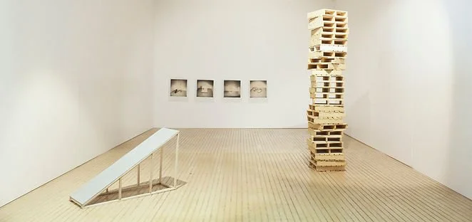 An art gallery with a tall wooden block sculpture, a ramp on the left, and four framed pictures on the back wall.