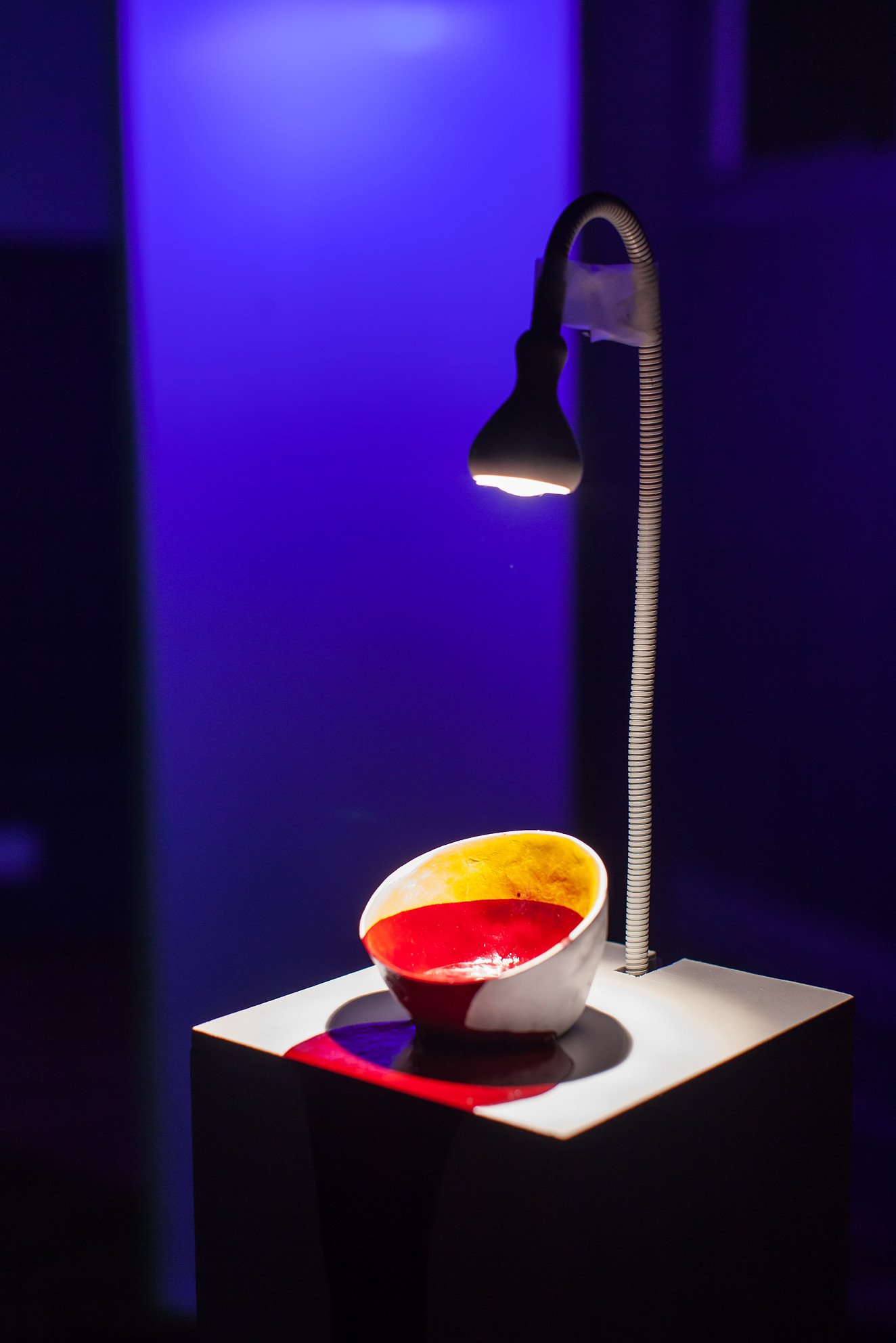 A ceramic bowl with red and yellow glaze under a small, flexible reading lamp on a pedestal in a dark room with blue lighting.