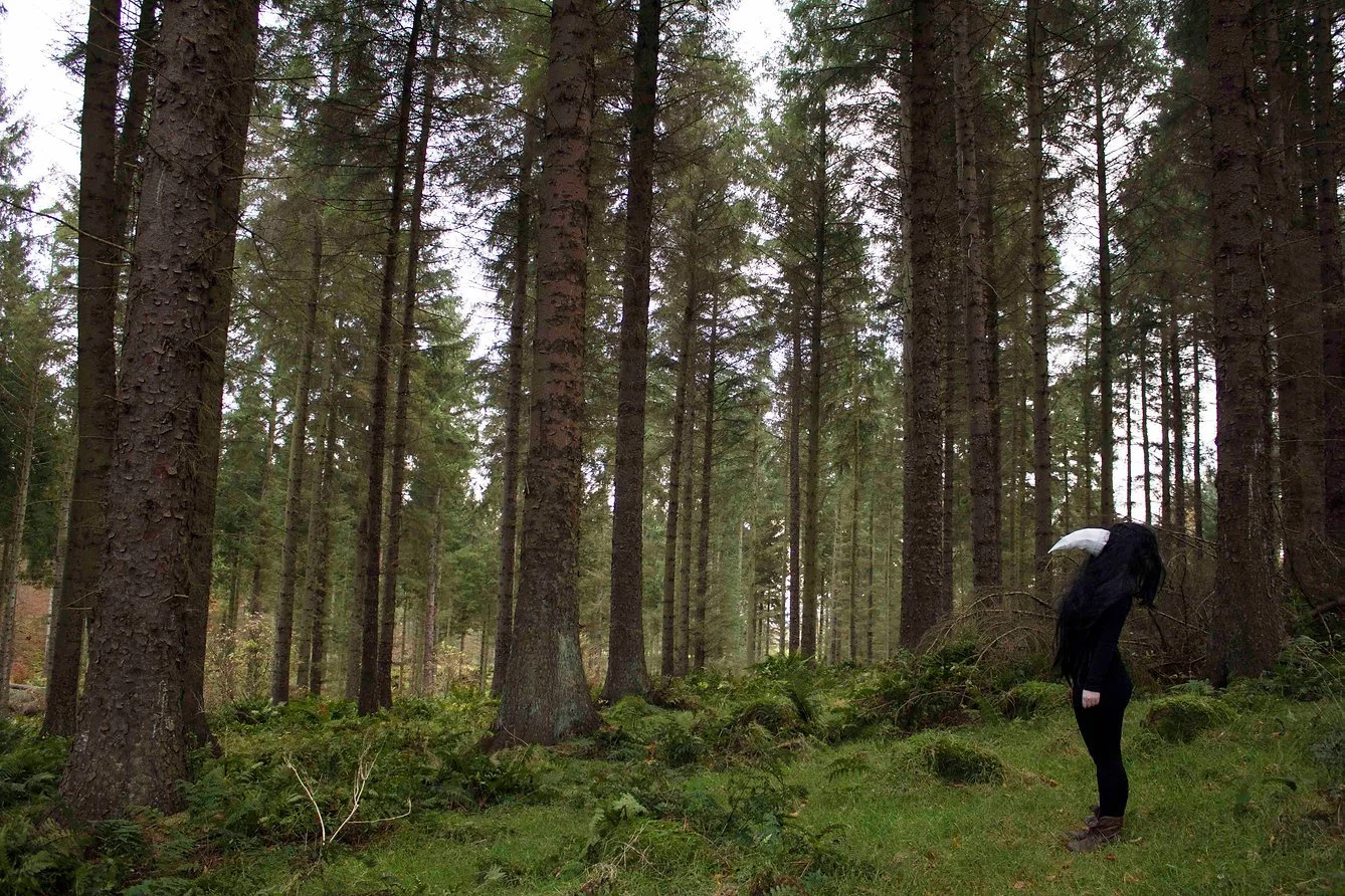A person dressed in black with long black hair and wearing a white bird beak, standing in a dense forest of tall trees with green foliage and moss on the ground.