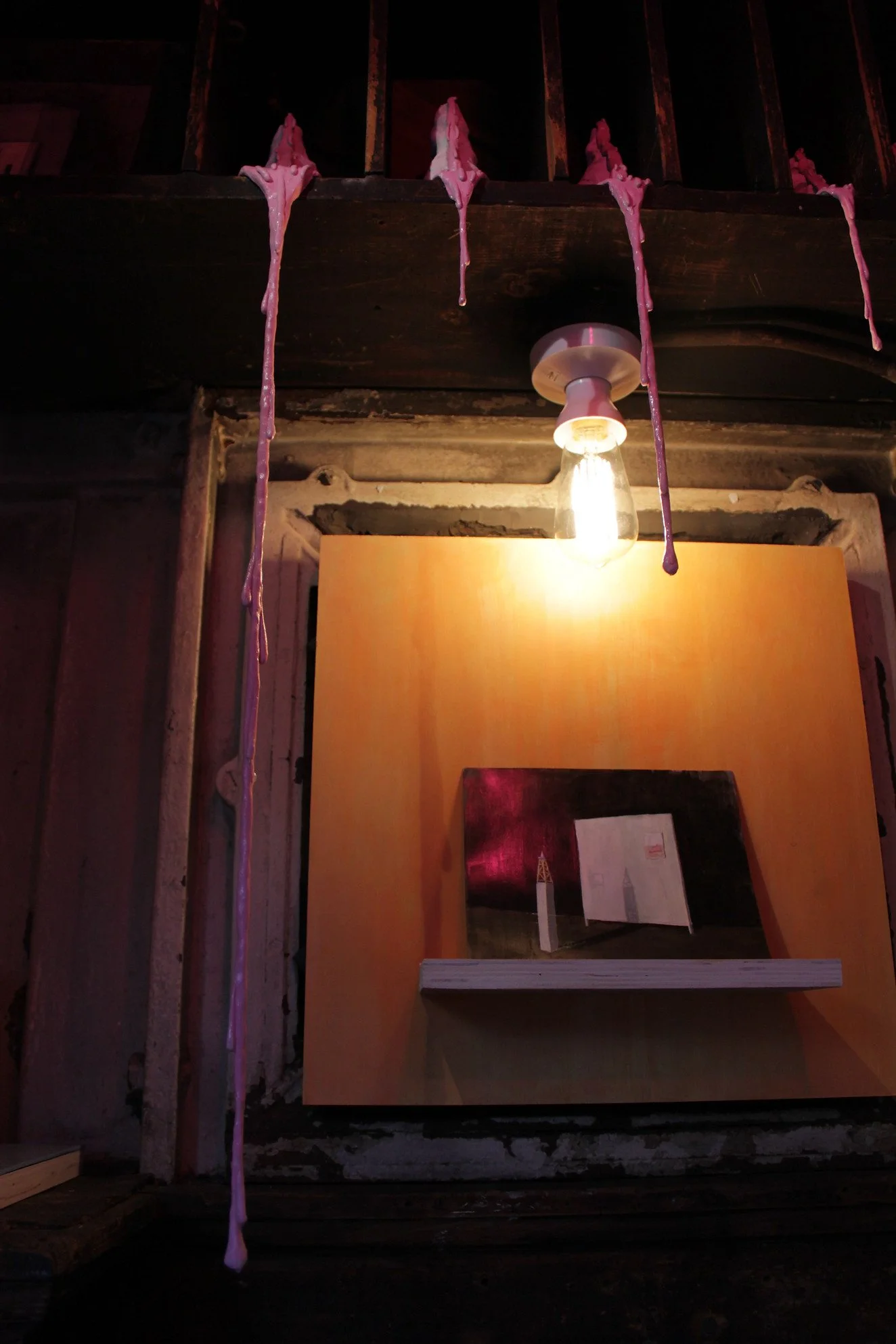 Pink wax drips from ceiling beams near a light bulb, with a reflection in a mirror on a yellow wall below.