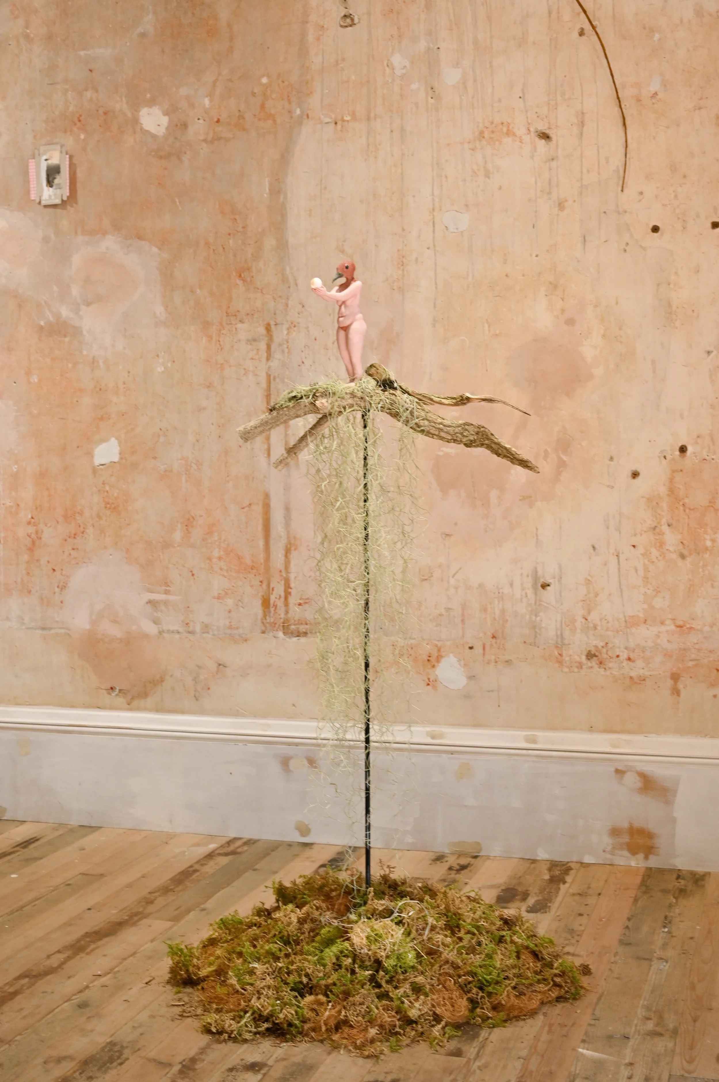 A sculpture of a person with a flamingo head, standing on a mossy mound and holding a ball, against a textured beige wall.