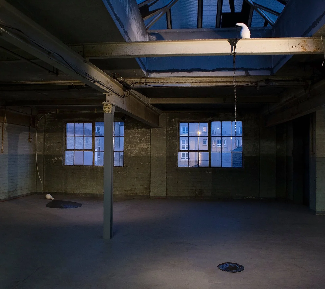 An empty industrial-style room with concrete floors and walls, two large windows showing neighboring buildings, a metal pole in the center, a ceiling with exposed piping, and a partially open skylight with a white ceramic pot perched on the edge.