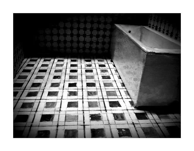 A black and white photo of a bathroom corner featuring a tiled floor with a square pattern, a cinder block bathtub, and patterned tile walls.