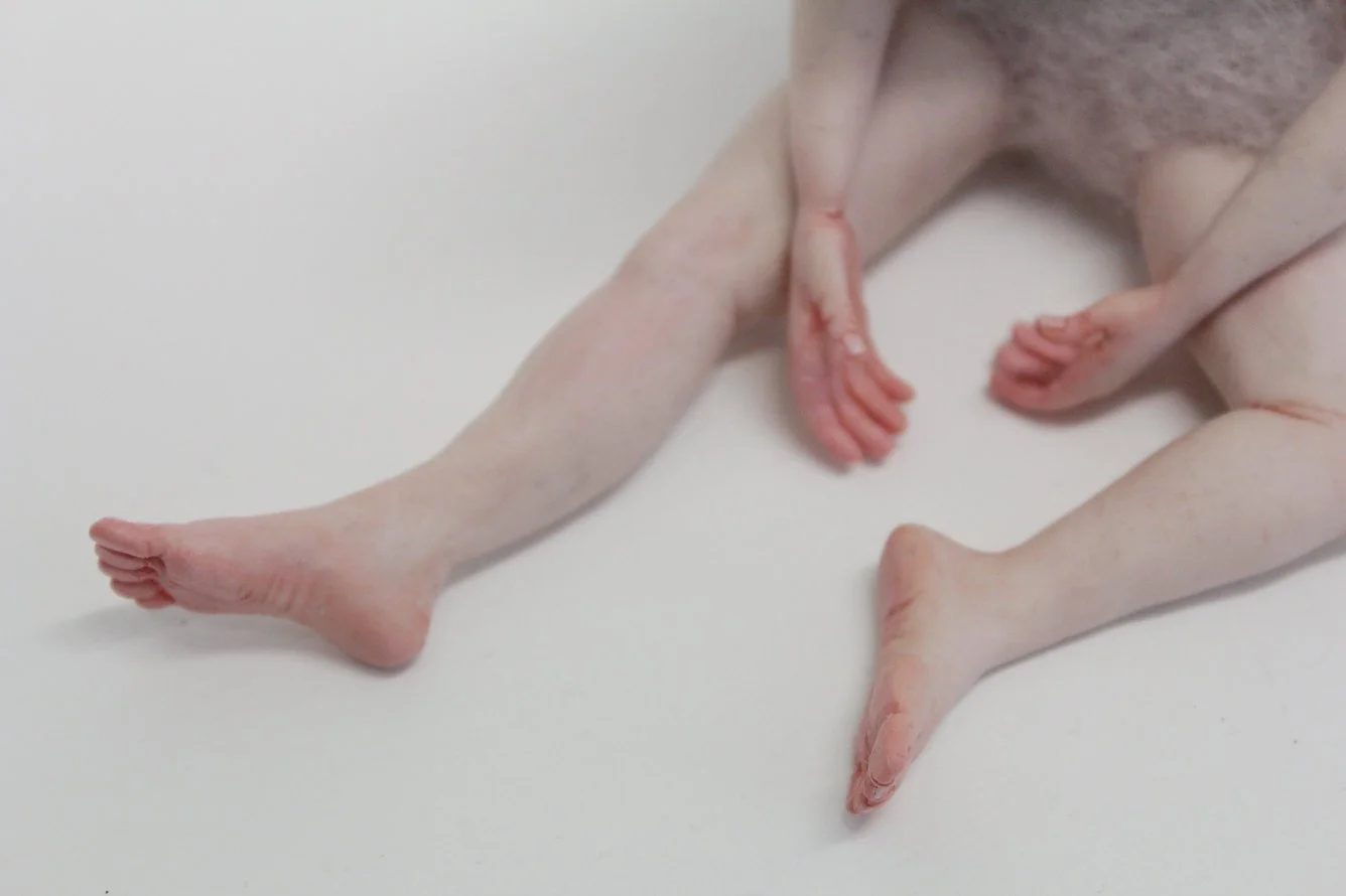 Close-up of a realistic sculpture showing legs, feet, and tiny hands, on a plain white background.