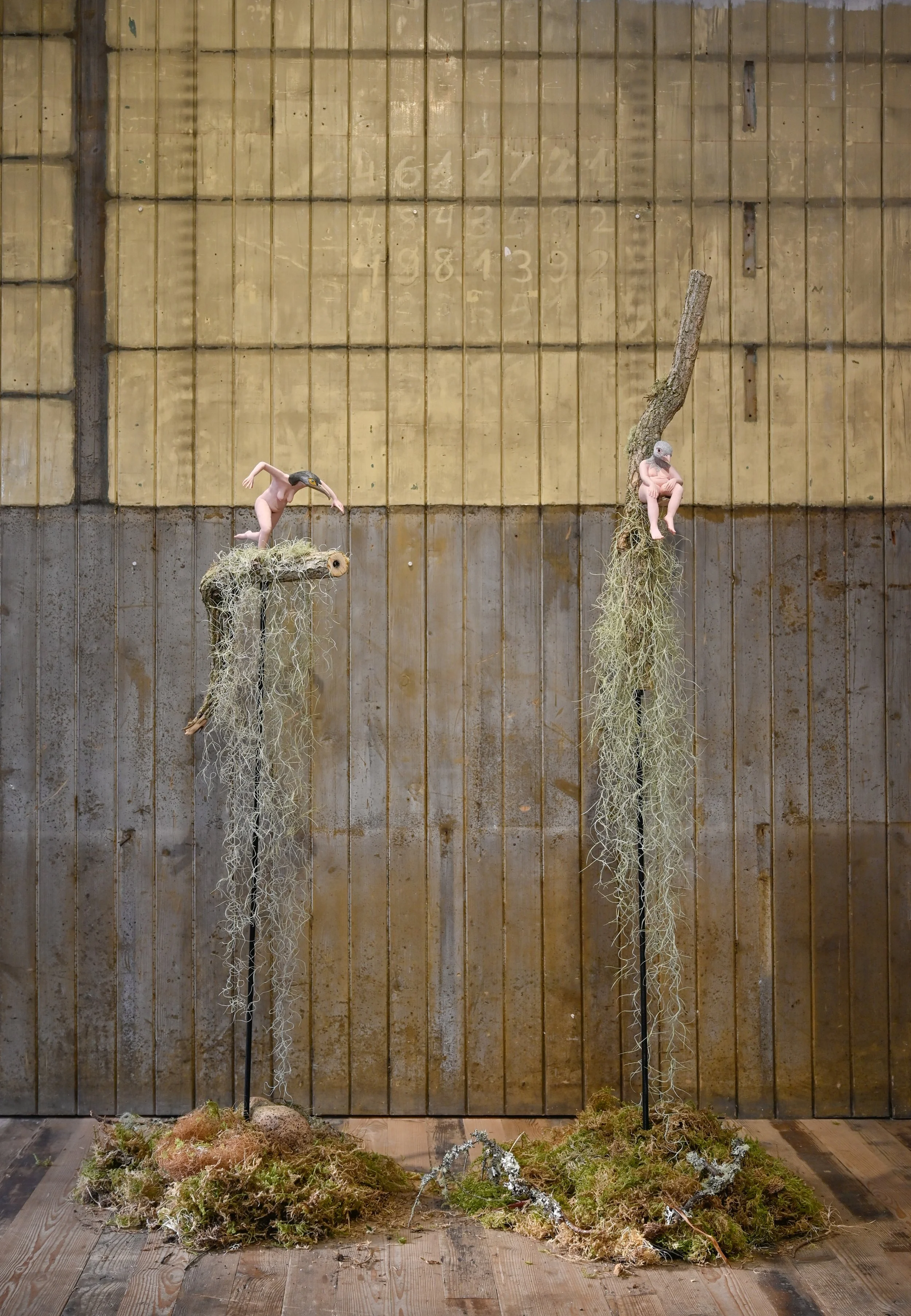 Two bird sculptures with human-like pink bodies and bird heads, perched on moss-covered stands against a wooden wall with chalk numbers faintly visible.